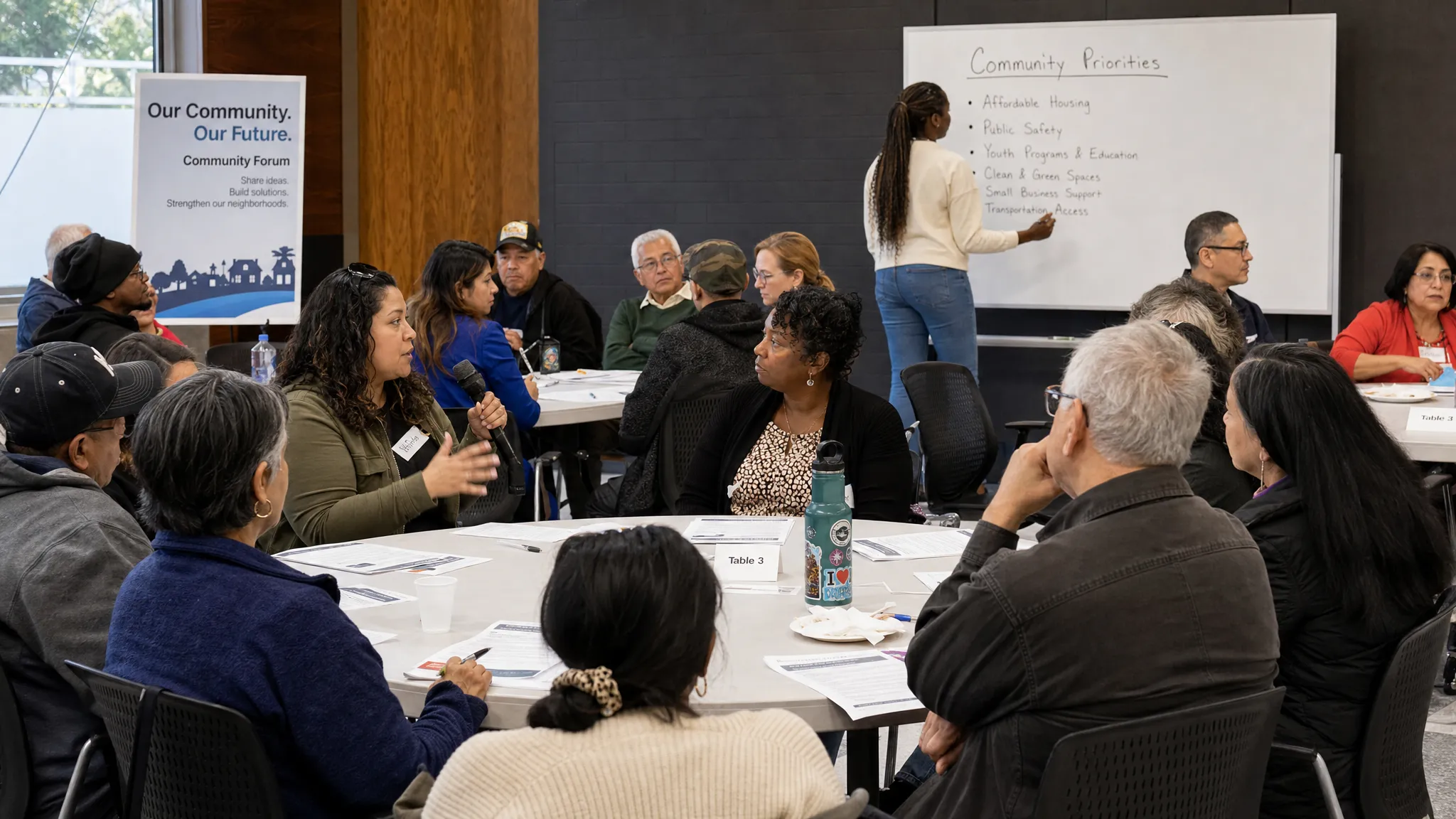 Adult residents gathered in a circular community forum with printed issue summaries on tables, listening to one another as a facilitator records shared priorities on a public board.
